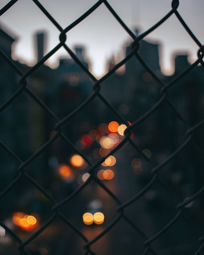 A chain-link fence at dusk with warm lights out of focus beyond it.