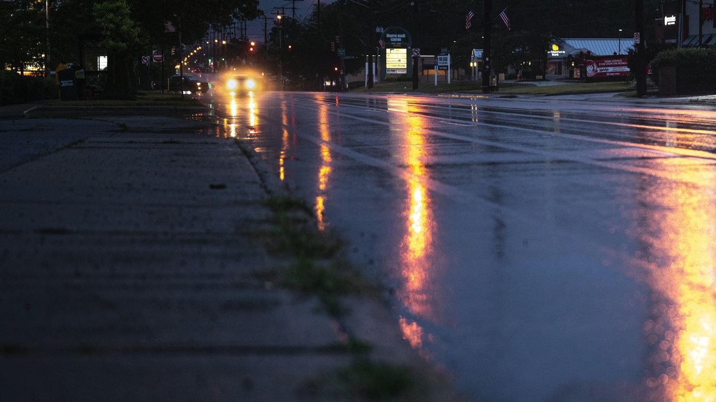 A wet road at dusk, headlights and sodium light reflecting off the pavement.